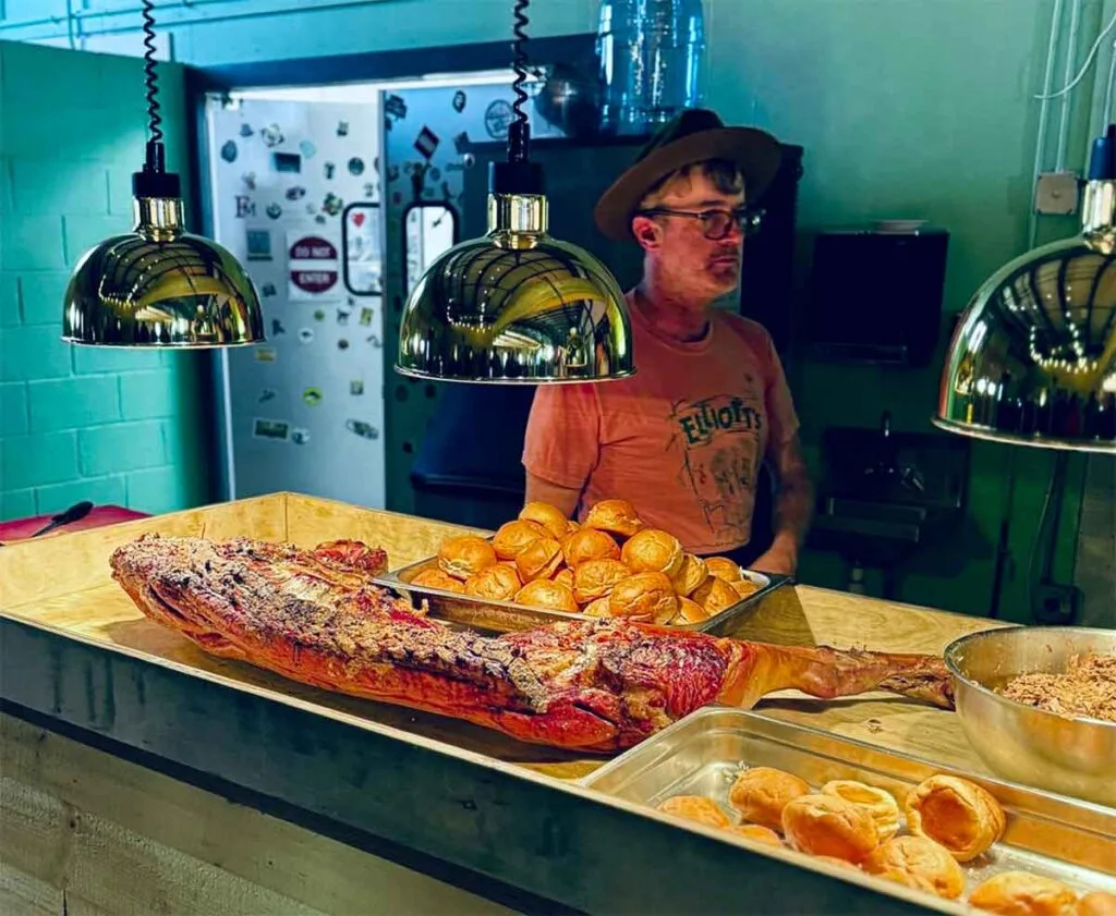 Elliott Moss behind service counter with half pig, buns, and pulled pork under hot lights.