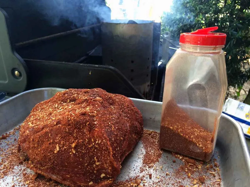 Pork shoulder coated in dry rub on a tray beside a rub shaker and a smoking charcoal chimney on the grill.