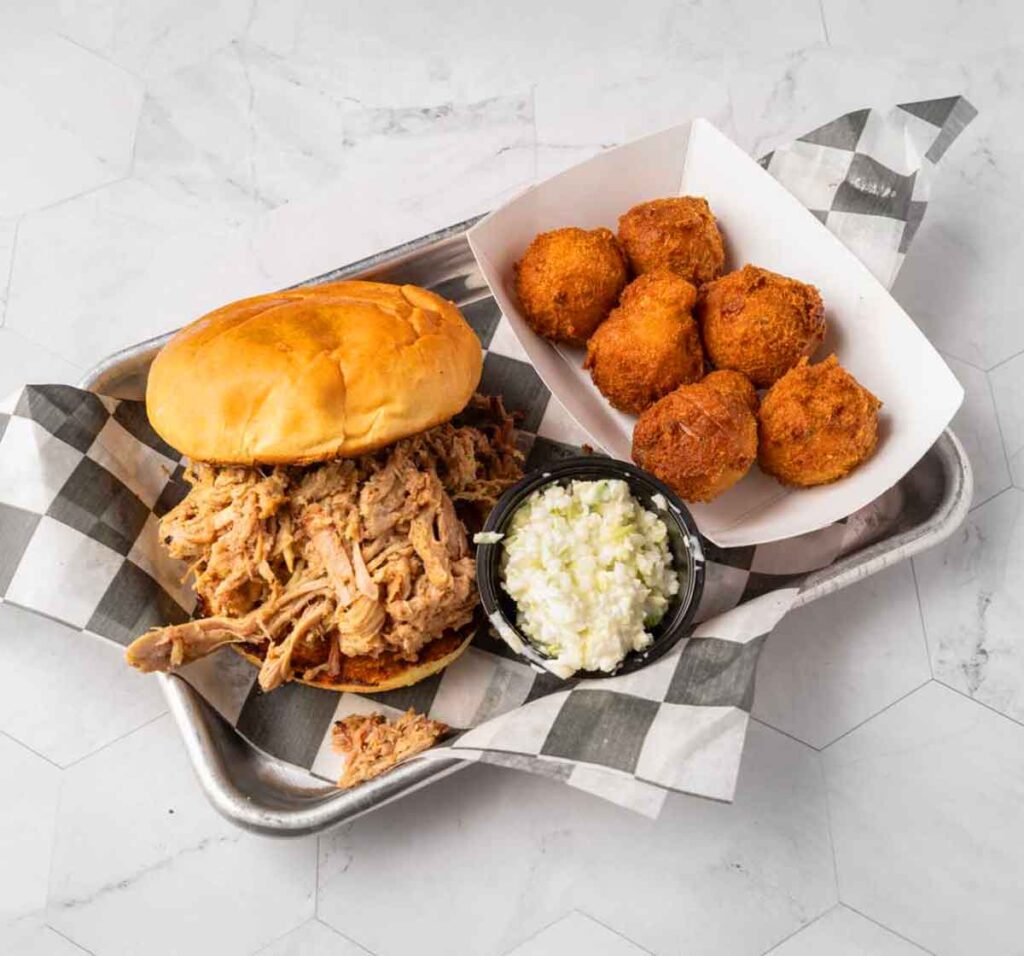 Pulled pork sandwich, slaw, and hush puppies on metal serving tray.