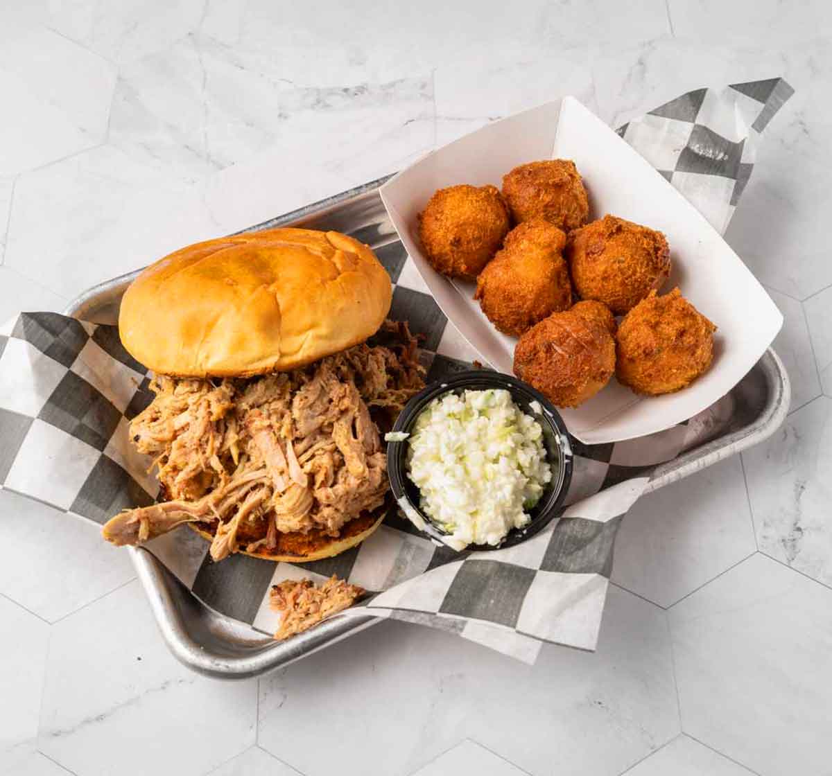 Pulled pork sandwich, slaw, and hush puppies on metal serving tray.