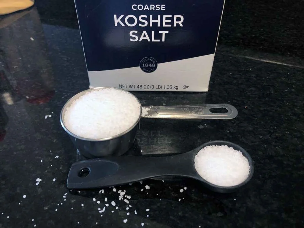Box of kosher salt on a black counter with a measuring cup and spoon filled with coarse salt crystals.