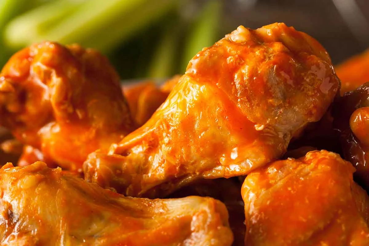 Close-up of buffalo wings coated in orange hot sauce, with celery blurred in the background.