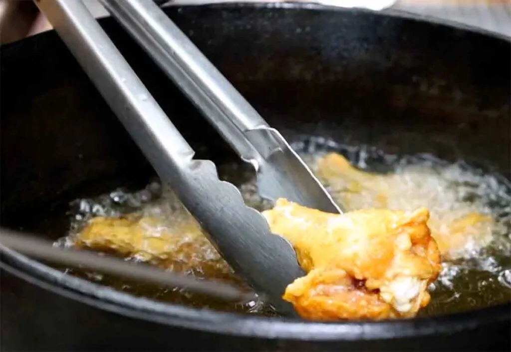Chicken wing frying in hot oil, lifted with tongs for a small-batch deep-fry workflow.