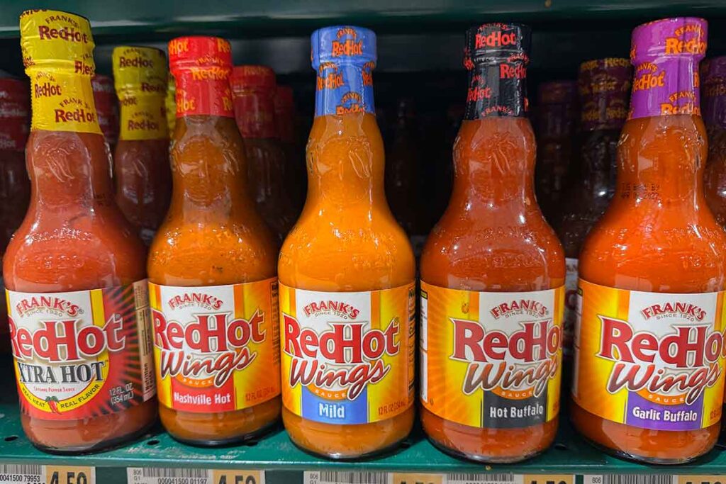 Frank’s RedHot Wing Sauce bottles on a store shelf, including Xtra Hot, Nashville Hot, Mild, Hot Buffalo, and Garlic Buffalo.