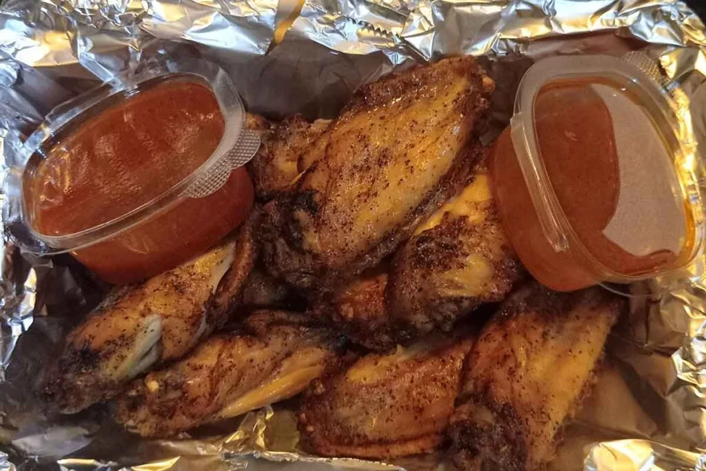 Dry-rubbed wings in a foil-lined tray with two cups of wing sauce on the side.