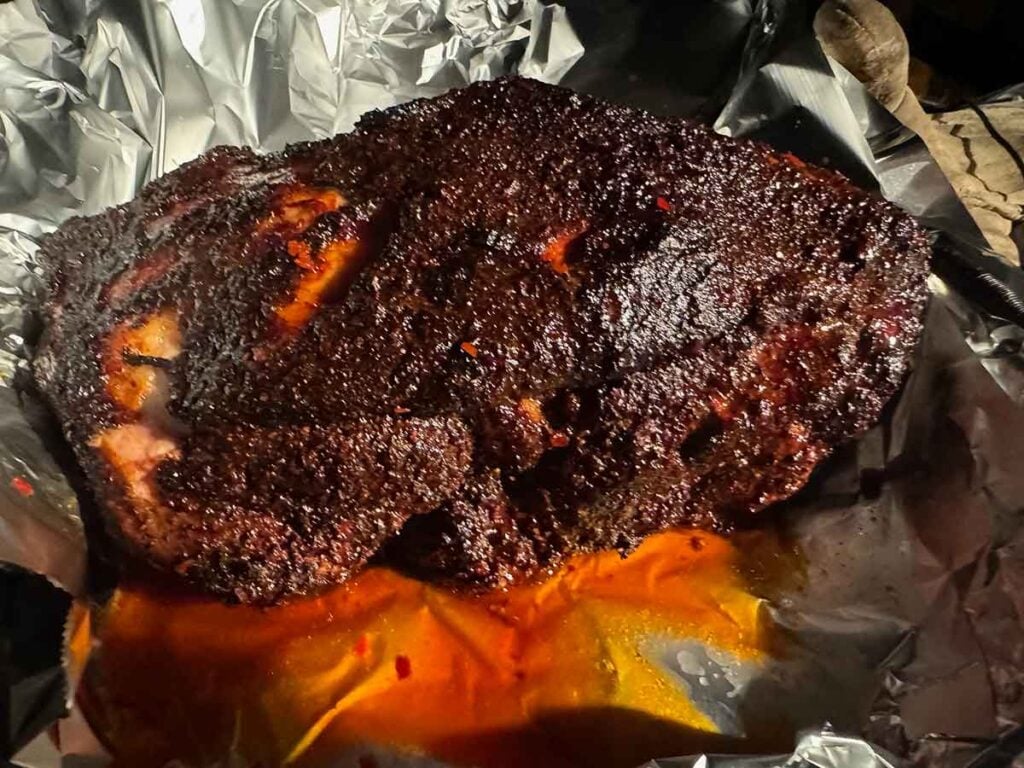 Smoked pork butt resting in opened foil, showing dark bark during the wrapping stage of the cook.