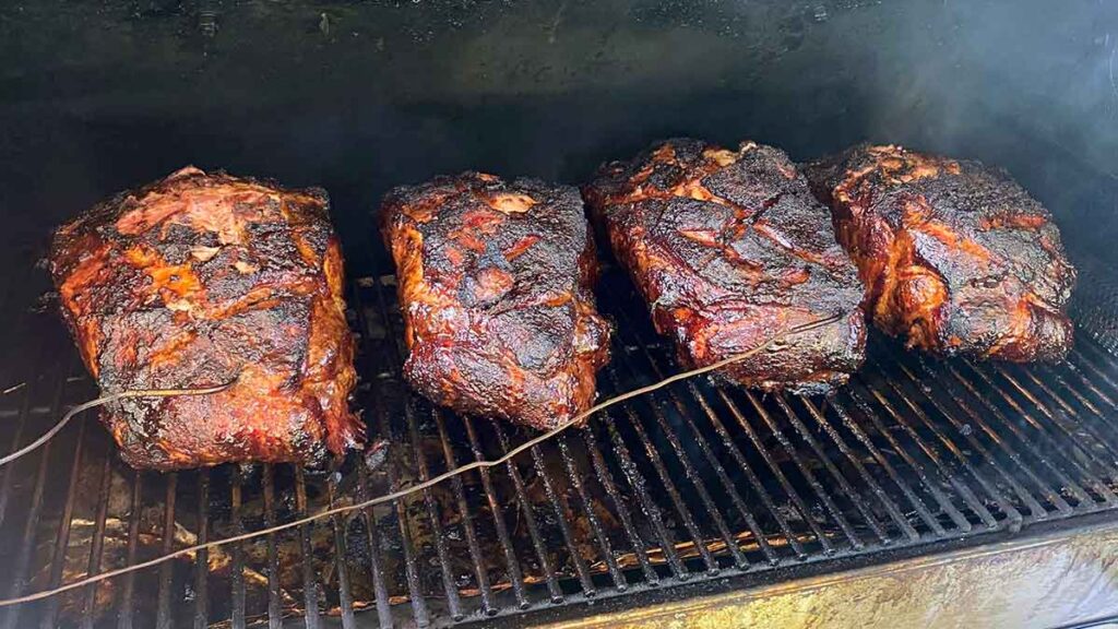 Four pork butts smoking side by side, illustrating how size, thickness, and shape can affect cook time.