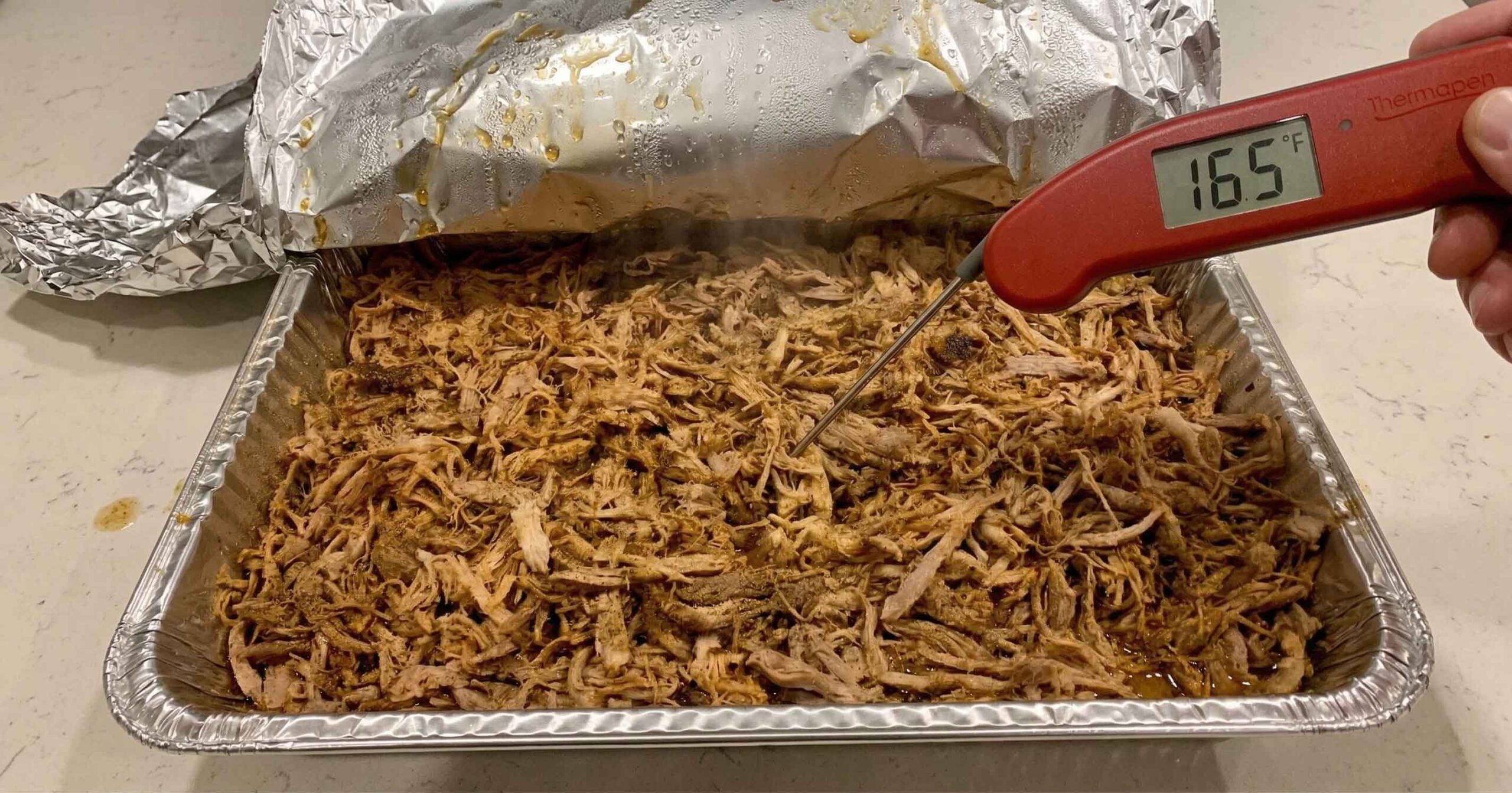 Reheated pulled pork in a foil-covered pan on a kitchen counter, checked with an instant-read thermometer at 165°F.