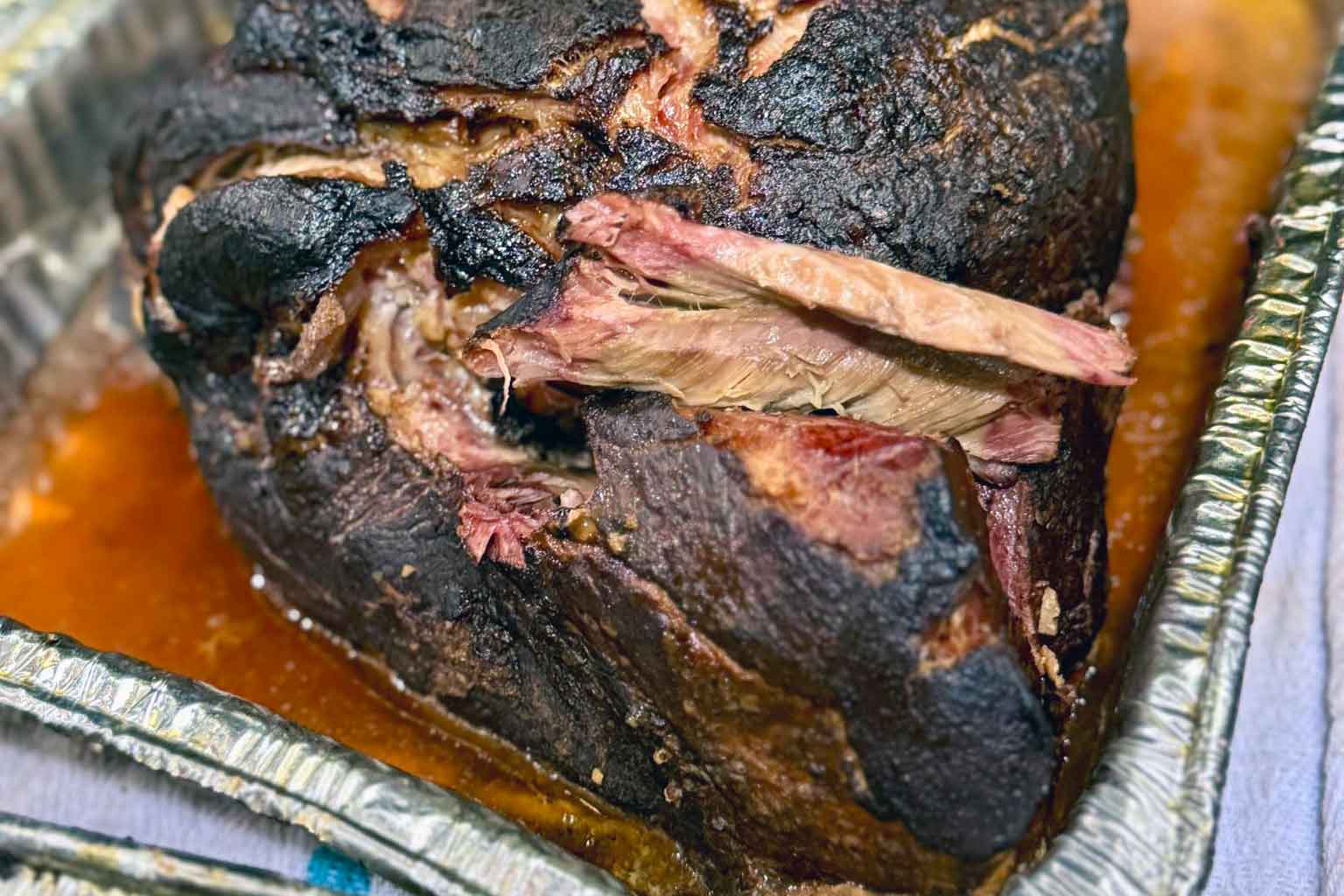 Whole smoked pork butt resting in a foil pan with reserved juices before being pulled for service.