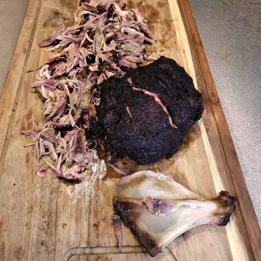 Pulled pork, a bark-covered section of smoked shoulder, and the removed shoulder bone on a cutting board after cooking.