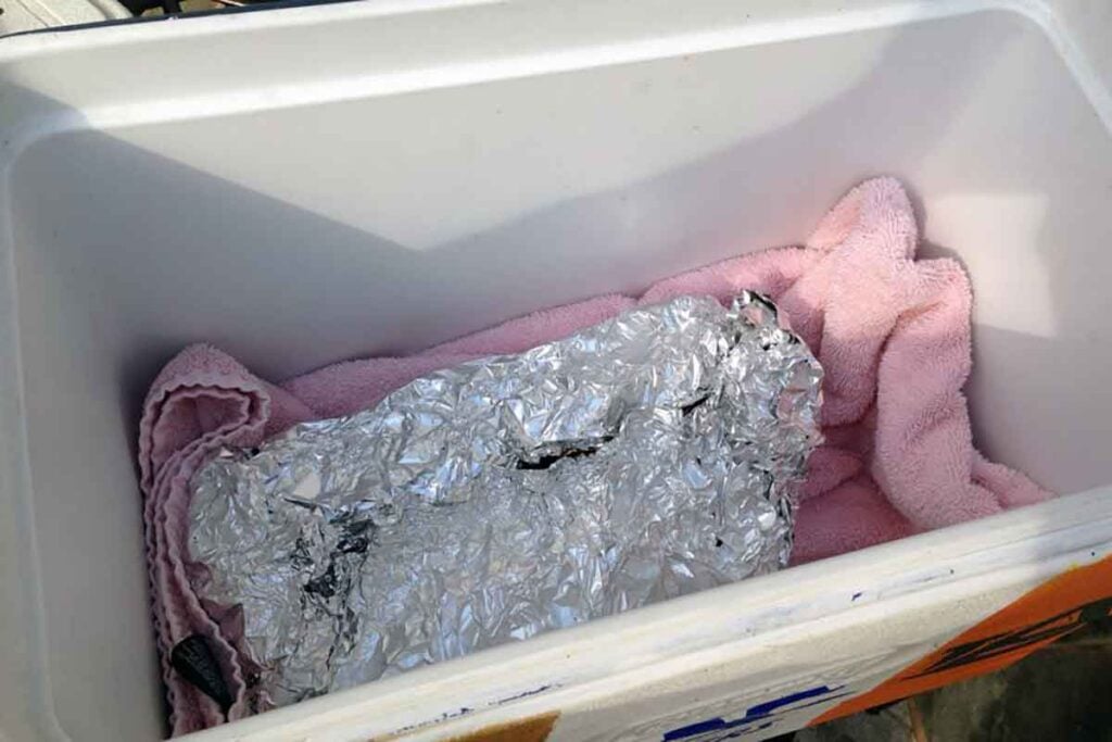 Foil-wrapped pork butt in a towel-lined cooler for holding pulled pork hot before serving.