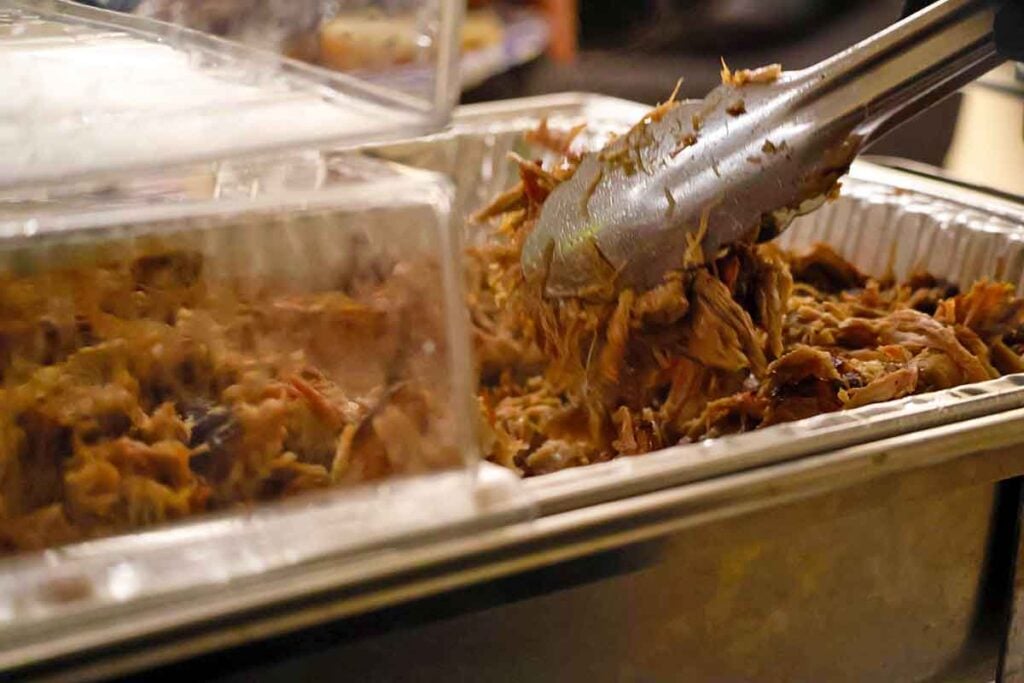Pulled pork held in a covered steam-table pan during service, with tongs lifting a portion from the batch.