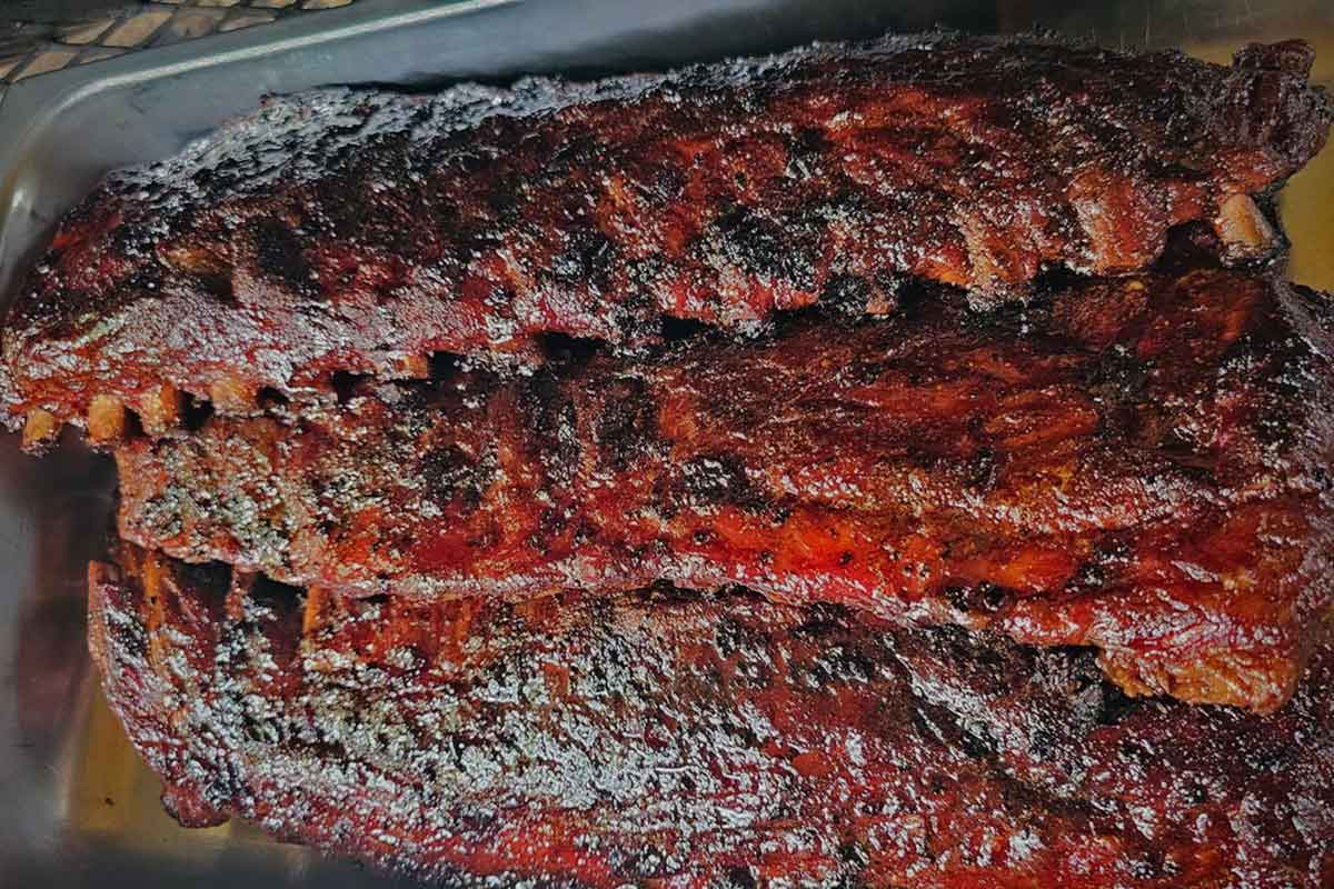 Whole racks of cooked pork ribs held in a pan before slicing and serving.