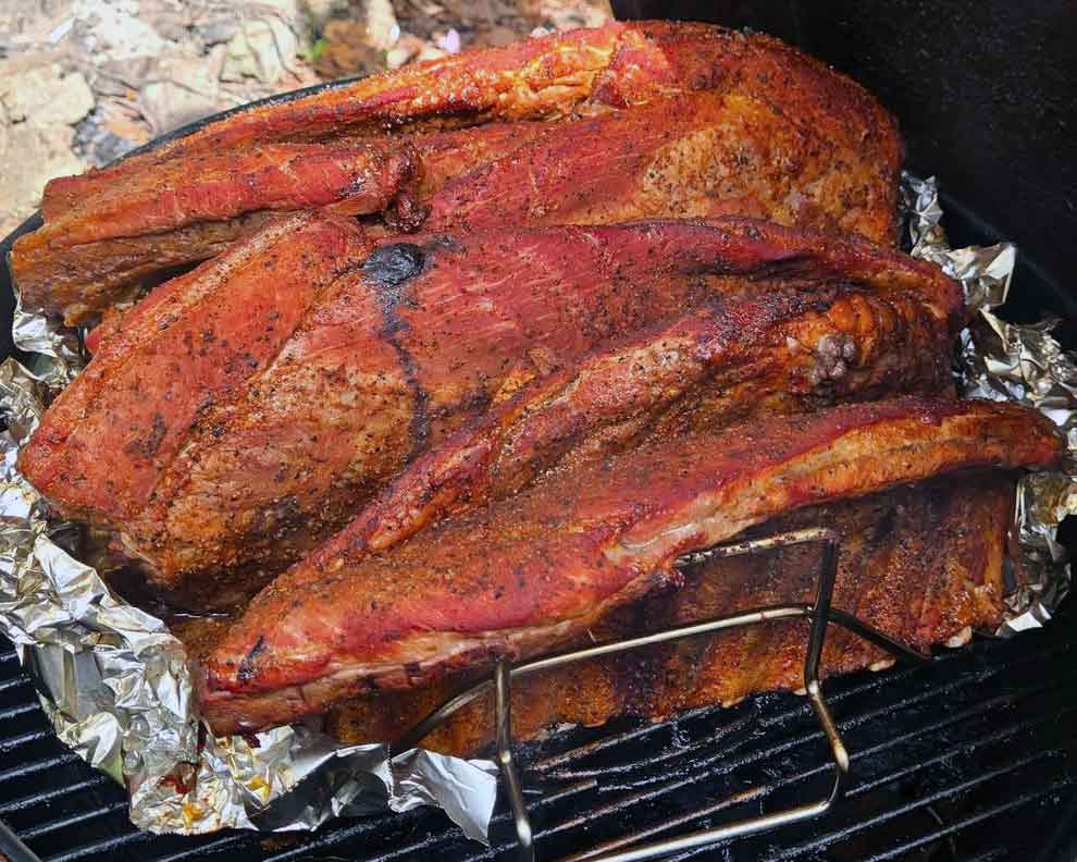 Several racks of ribs crowded upright in a rib holder on a small charcoal grill.