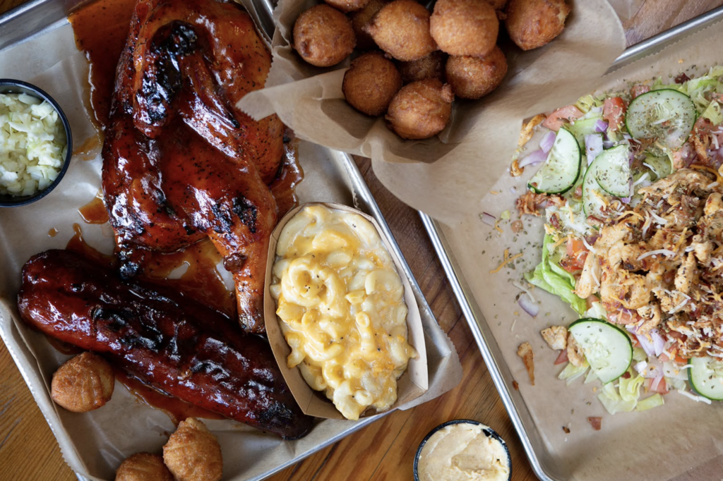 Tray with smoked meats covered in sauce with sides beside a basket of hush puppies and another tray with a salad.