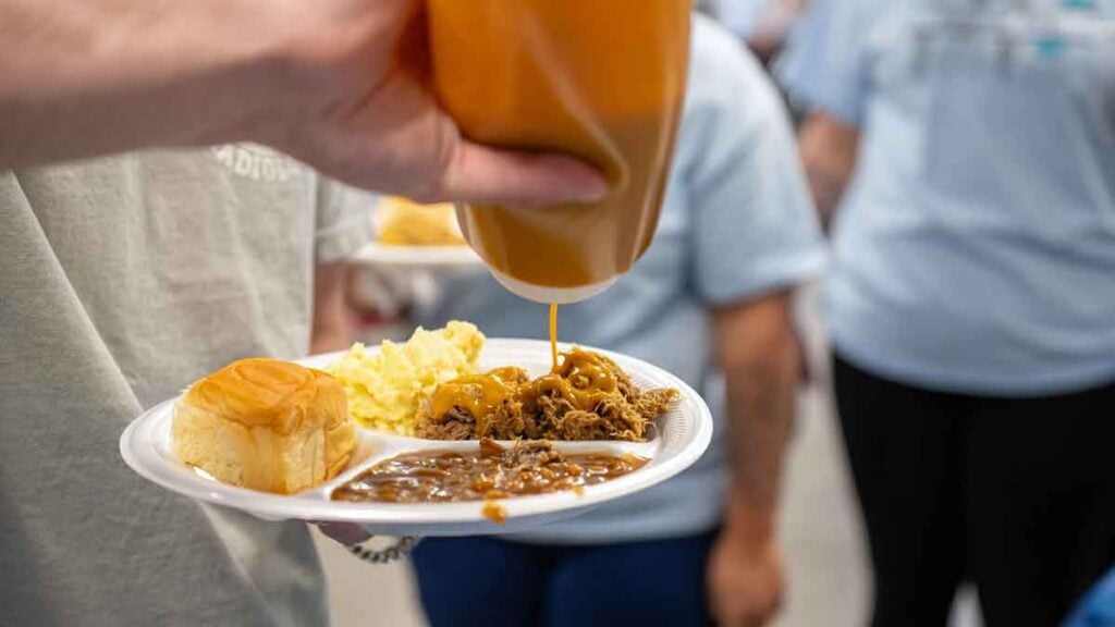 Hand squeezing mustard BBQ sauce onto a plate holding pulled pork and sides.